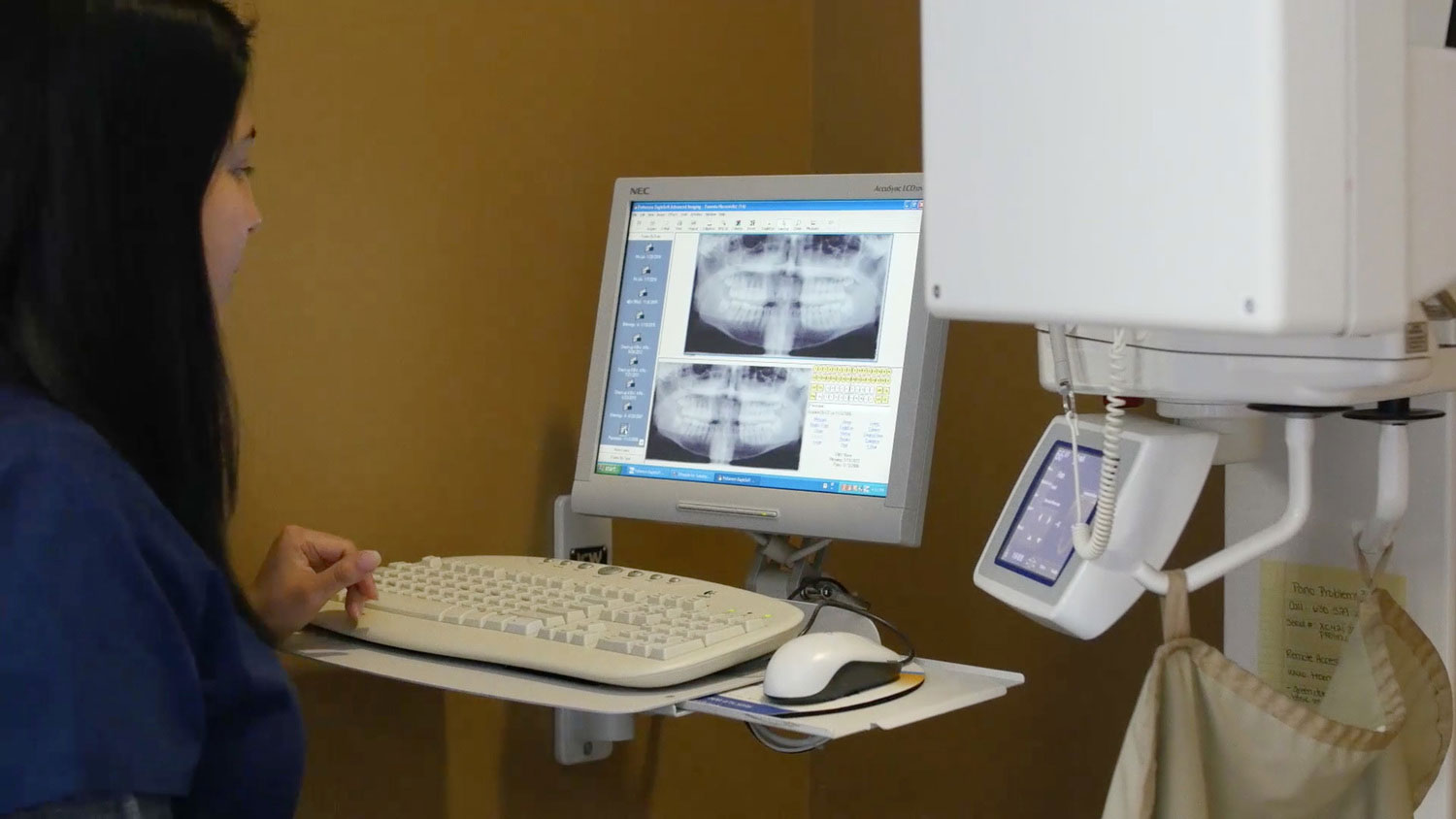 A dental hygienist standing at a computer monitor displaying an X-ray image of a patient s teeth, with a keyboard and mouse nearby.