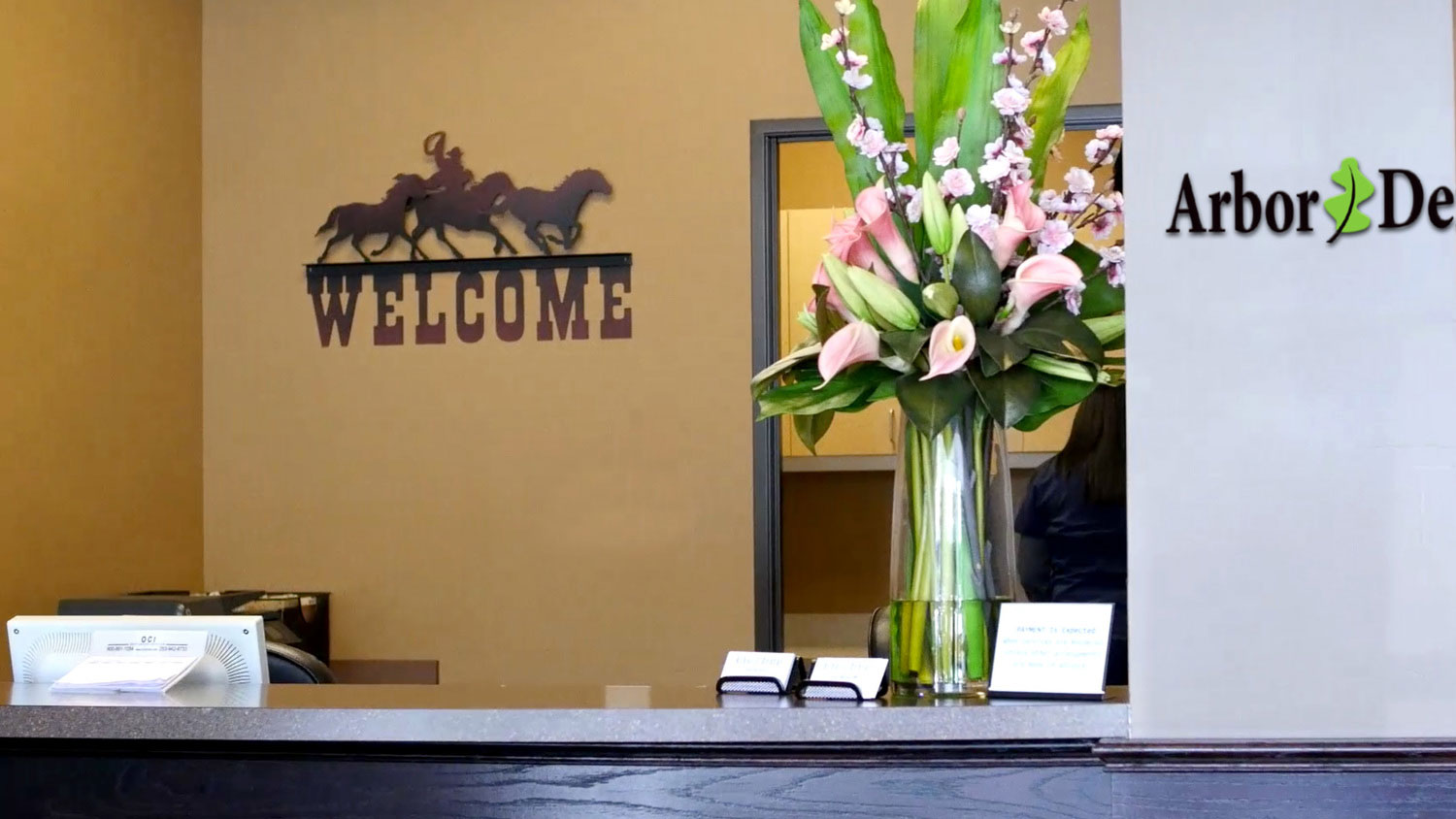 The image shows an interior view of a reception area with a sign that reads Welcome above a desk, and there s a wall decoration with the text Arbor De La Costa.