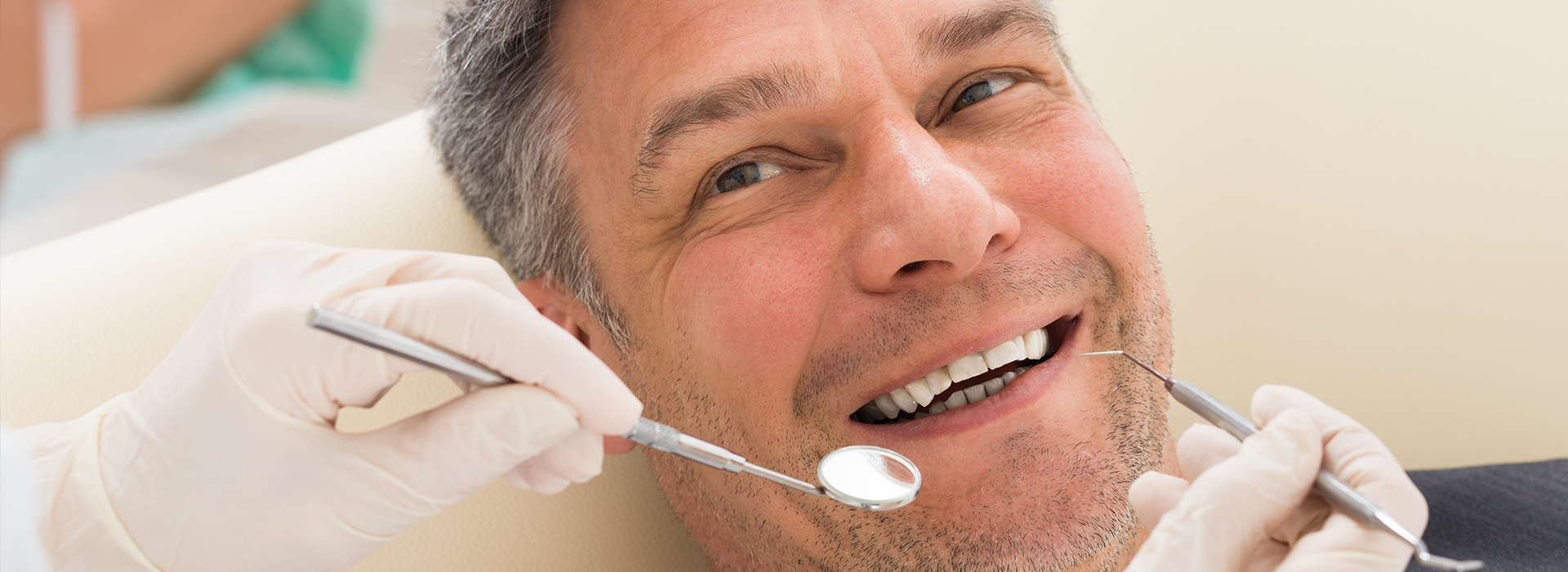The image shows a man sitting in a dental chair with a smiling expression, receiving dental treatment from a dentist who is working on his teeth.