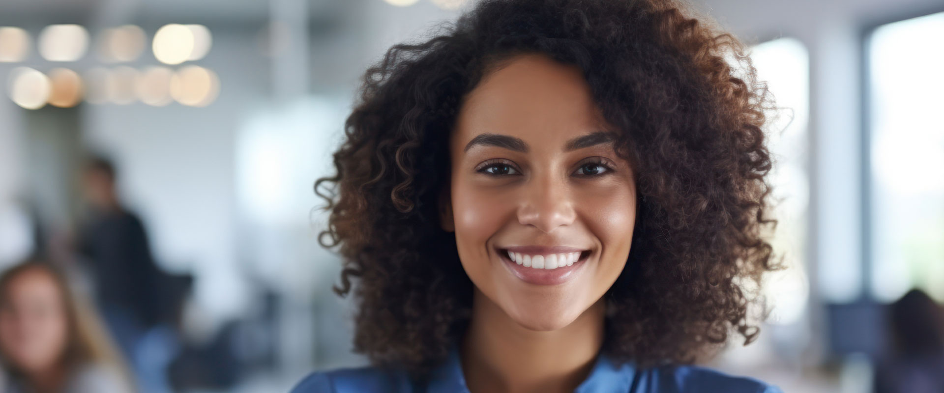The image shows a professional woman with curly hair, smiling at the camera, wearing business attire, standing indoors with blurred background elements that suggest an office environment.